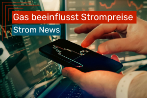 Ein Smartphone wird in den Händen gehalten, auf dem ein Kursverlauf angezeigt wird, während im Hintergrund ein Computerbildschirm mit weiteren Finanz- oder Energiediagrammen zu sehen ist.